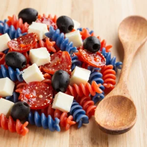 A vibrant red, white, and blue pasta salad served in a clear bowl, featuring cherry tomatoes, mozzarella balls, and blue cheese.